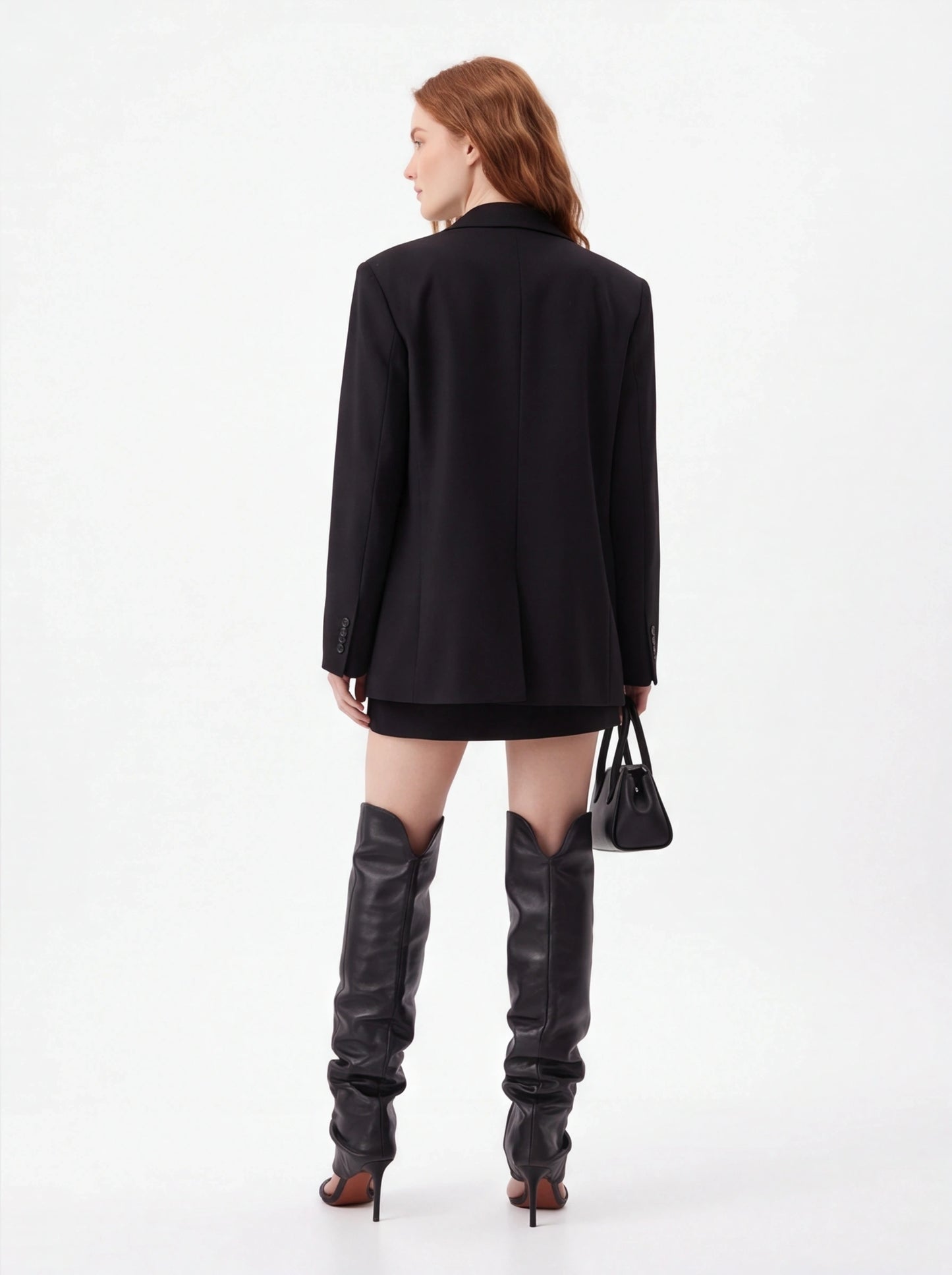 A model shown from the back wearing an oversized black blazer, a short black skirt, and tall black leather over-the-knee boots, holding a small black handbag against a plain white studio background.