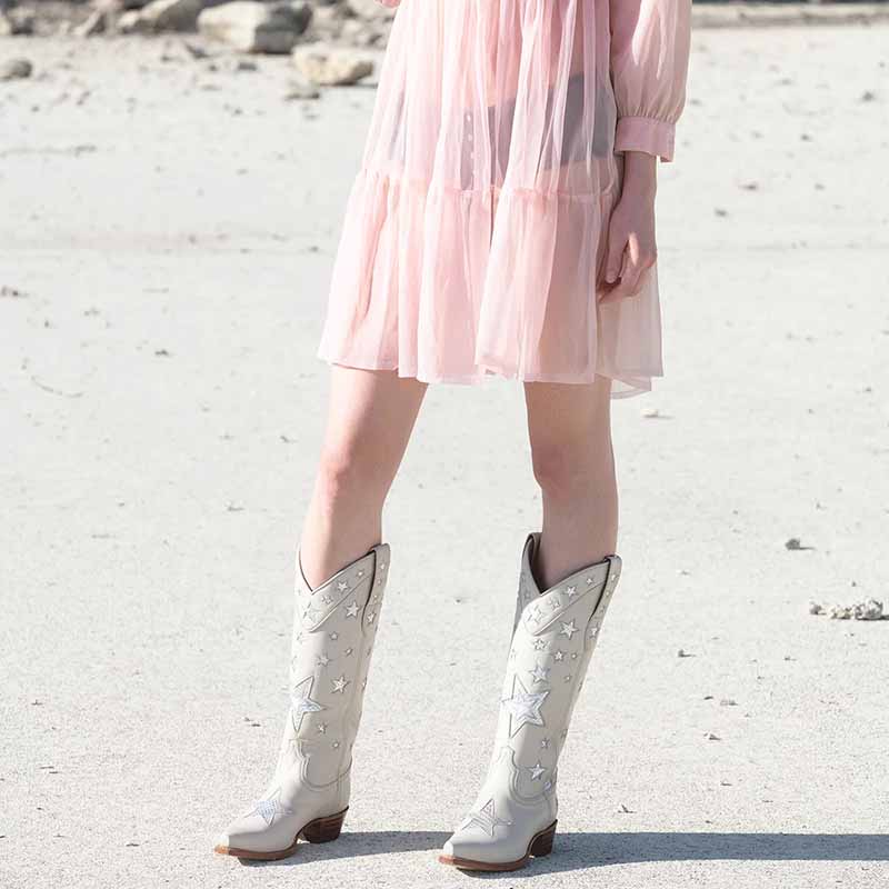 Star embroidered cowboy boots in white with silver stars, styled with a pink dress on a beach