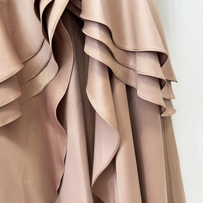 Close-up of a draped garment in a soft taupe-beige satin-like fabric with layered ruffles and flowing folds against a plain light background.