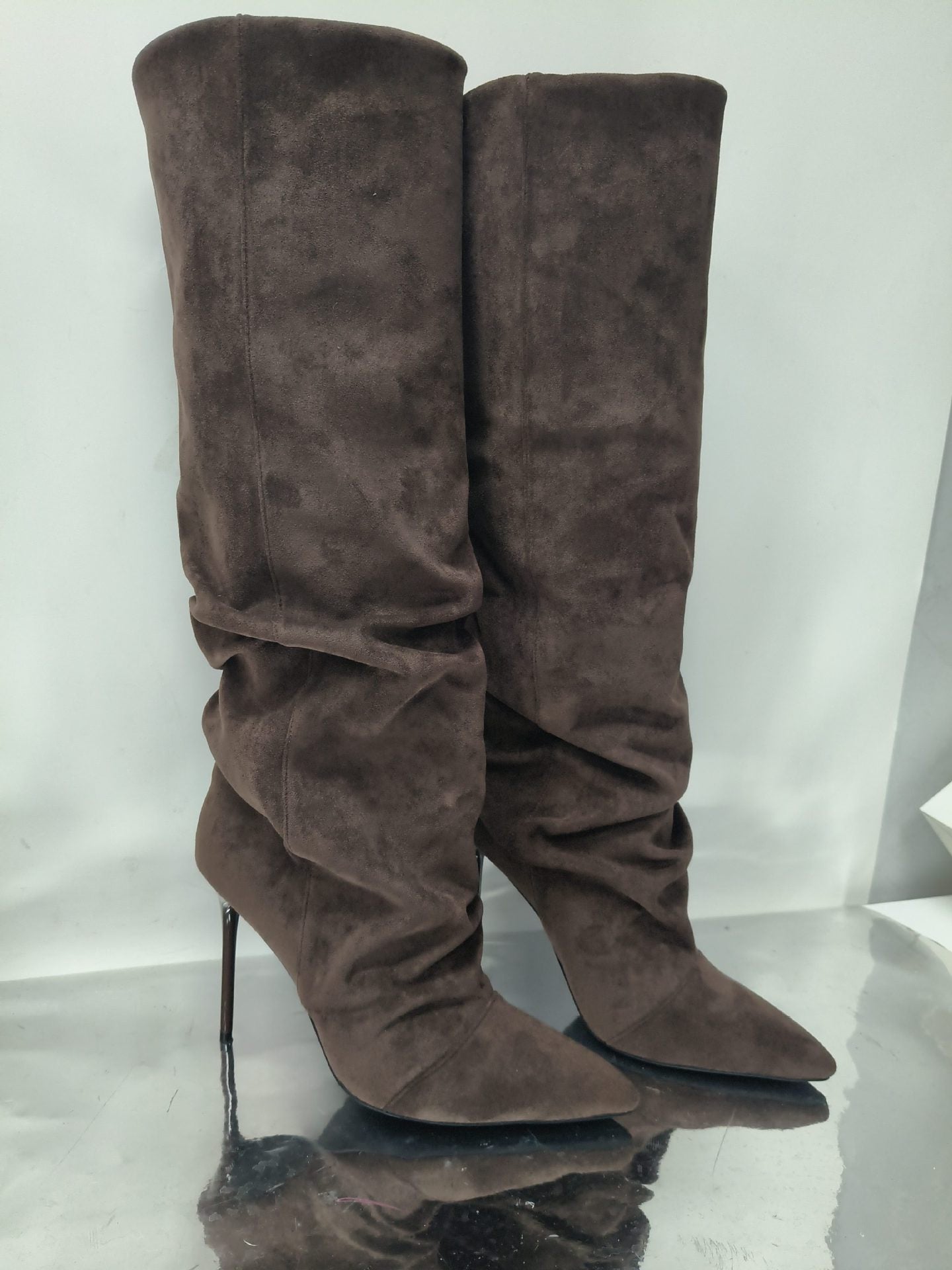 A pair of tall brown suede knee-high boots with pointed toes and slim high heels, standing on a reflective surface against a light gray background.