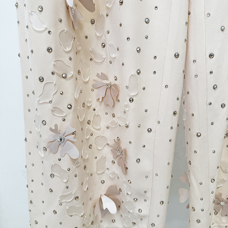 A close-up of a cream-colored fabric garment featuring 3D floral appliqués in shades of beige and white, embellished with scattered silver rhinestones.