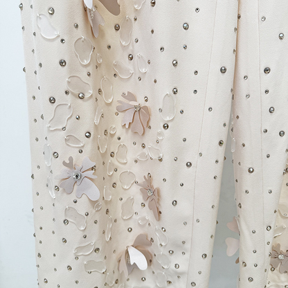 A close-up of a cream-colored fabric garment featuring 3D floral appliqués in shades of beige and white, embellished with scattered silver rhinestones.