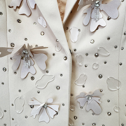 Close-up of a cream-colored jacket or blazer with decorative white fabric flowers, clear rhinestone accents, and small silver studs.