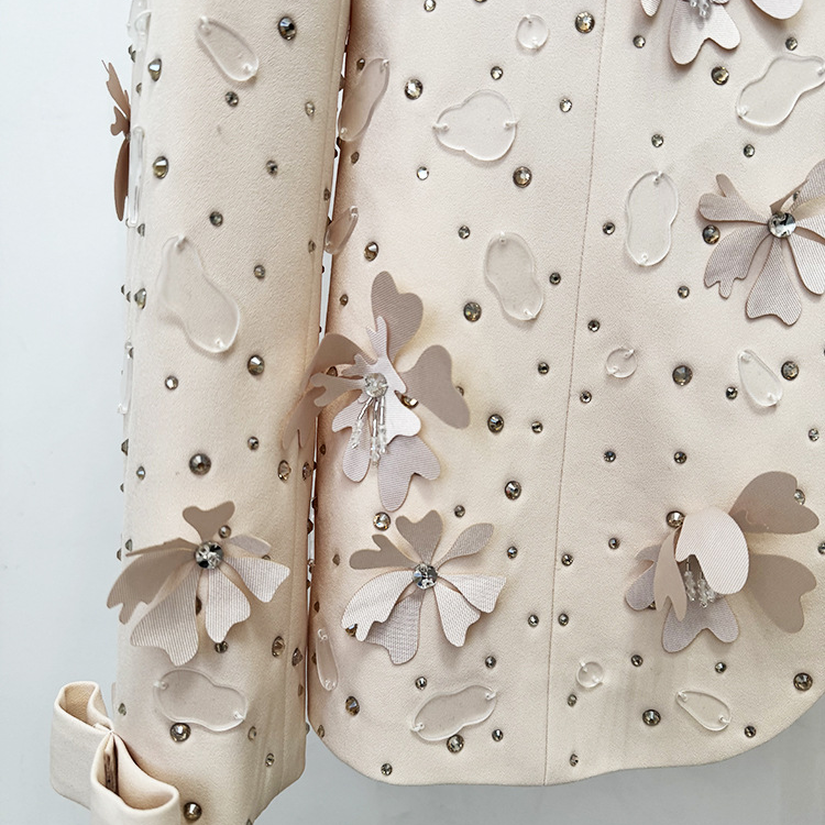 A close-up of a beige embellished jacket or coat with 3D floral appliqués, rhinestone studs, and a matching rolled sleeve, shown against a plain light background.