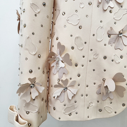 A close-up of a beige embellished jacket or coat with 3D floral appliqués, rhinestone studs, and a matching rolled sleeve, shown against a plain light background.