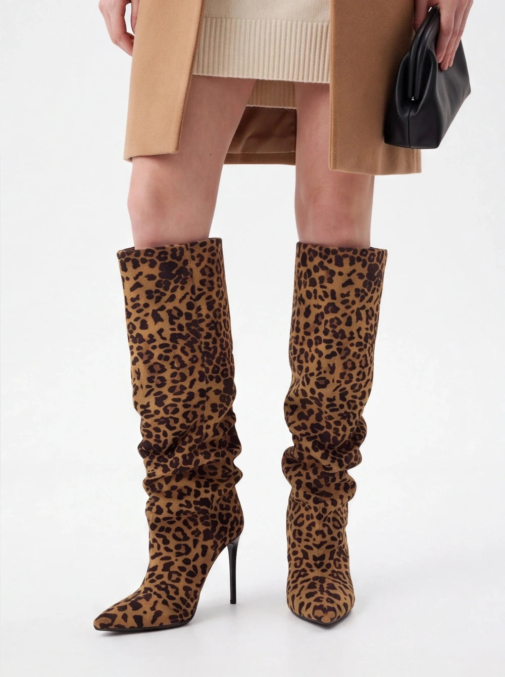 A person wearing knee-high leopard-print heeled boots with a beige ribbed knit top, tan coat, and black handbag against a light gray studio background.