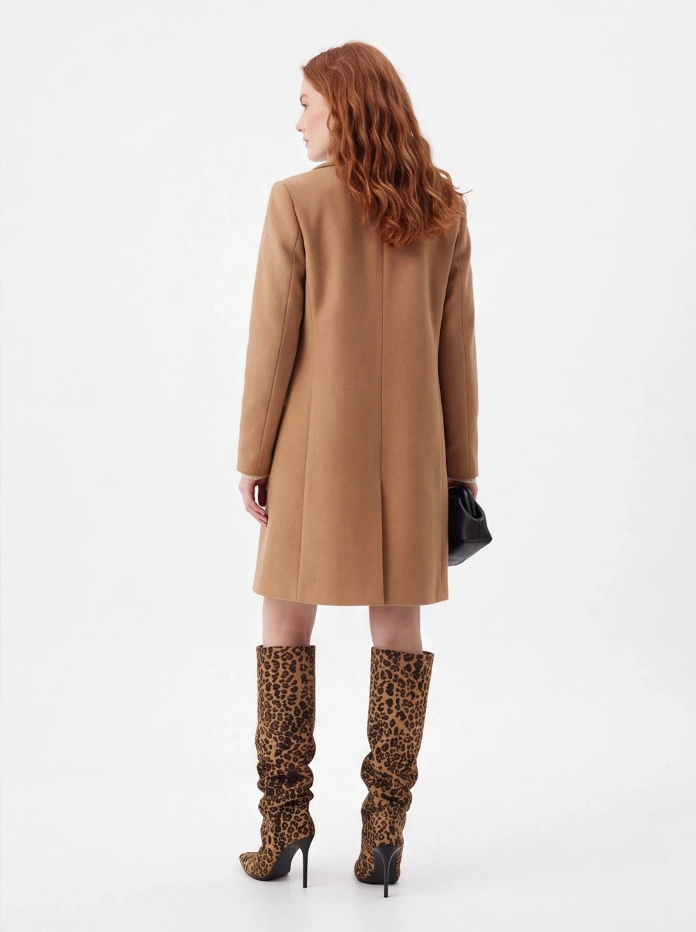 A woman with wavy red hair is shown from the back wearing a camel-colored knee-length coat and leopard-print knee-high heeled boots, holding a small black clutch against a plain white studio background.
