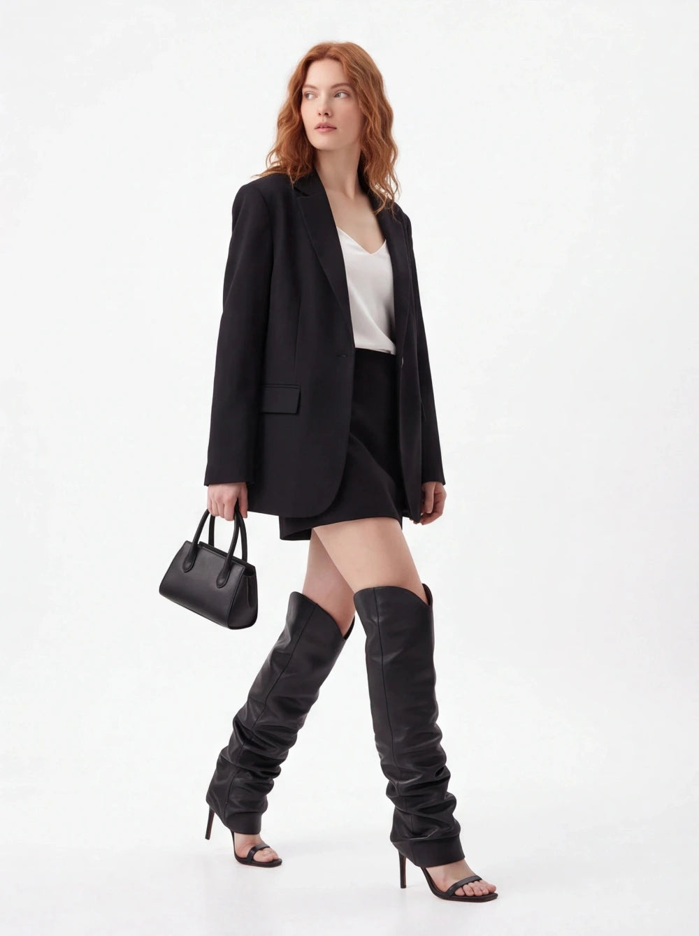 A woman models a black blazer, black mini skirt, white top, black thigh-high slouchy boots, and a black handbag against a plain white studio background.