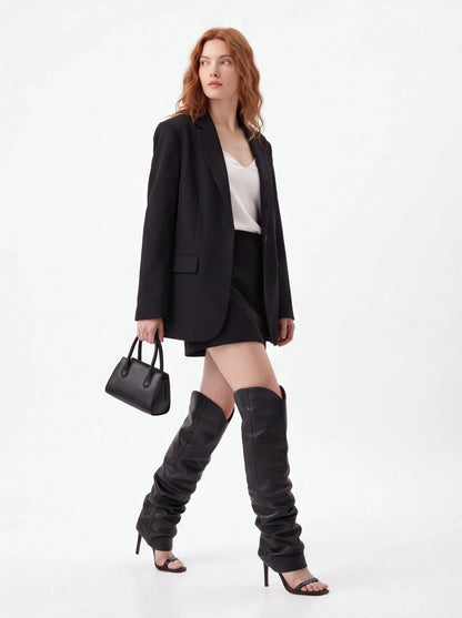 A woman models a black blazer, black mini skirt, white top, black thigh-high slouchy boots, and a black handbag against a plain white studio background.