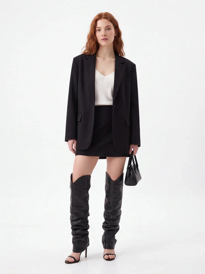 A model stands against a plain white studio background wearing an oversized black blazer, black mini skirt, black thigh-high slouchy boots, a white top, and black heeled sandals, while holding a small black handbag.
