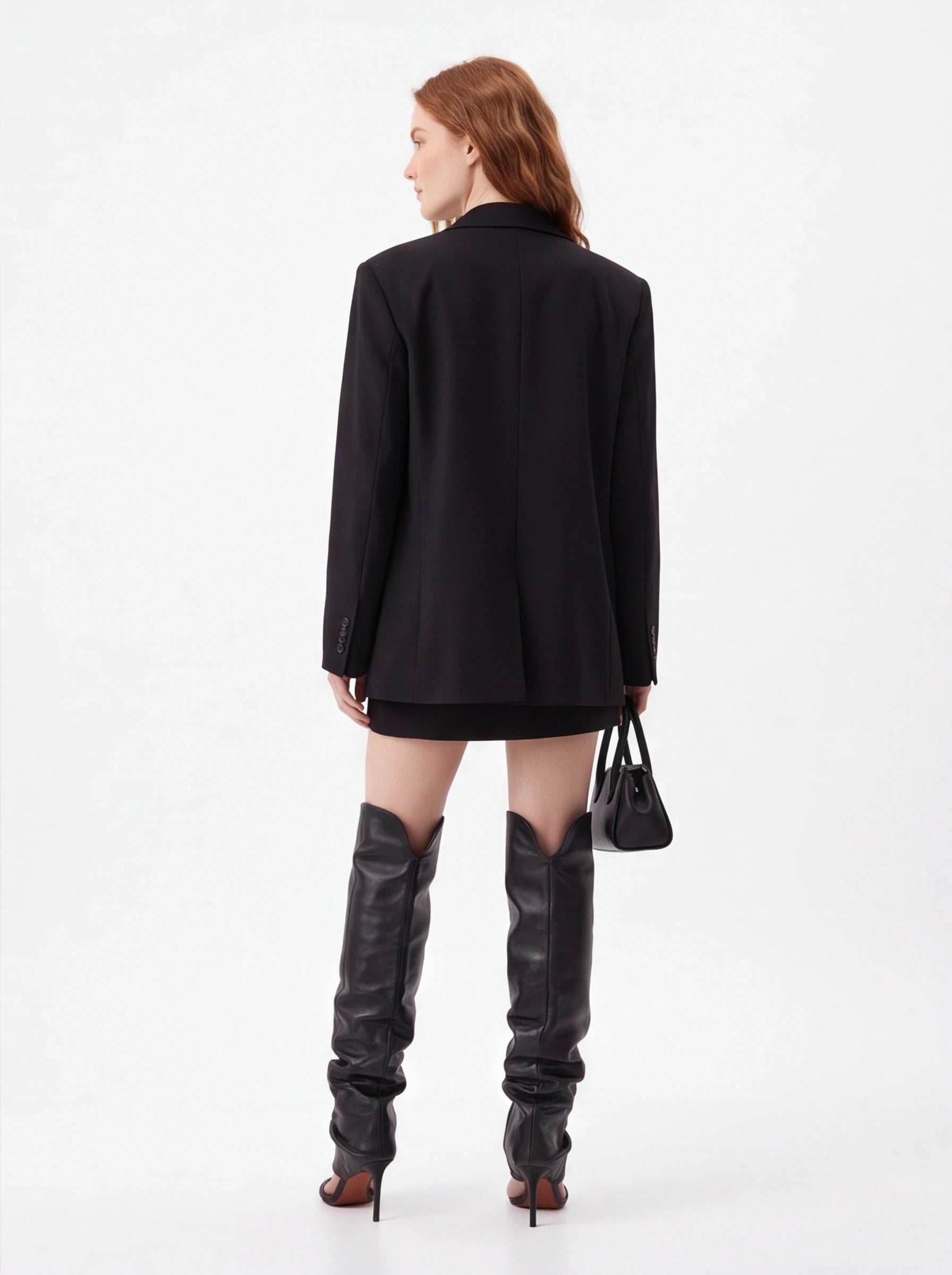 A model shown from the back wearing an oversized black blazer, a short black skirt, and tall black leather over-the-knee boots, holding a small black handbag against a plain white studio background.
