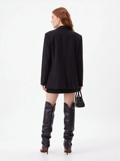 A model shown from the back wearing an oversized black blazer, a short black skirt, and tall black leather over-the-knee boots, holding a small black handbag against a plain white studio background.