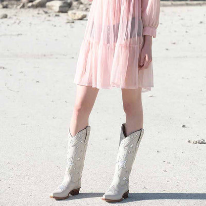 Star embroidered cowboy boots in white with silver stars, styled with a pink dress on a beach