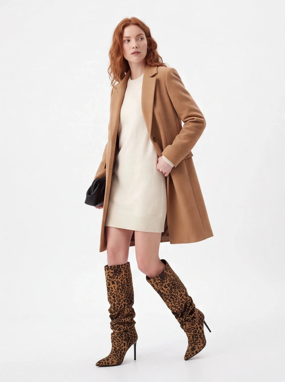A model wearing a camel mid-length coat over a cream mini dress, paired with leopard-print slouchy knee-high boots and holding a small black clutch, photographed against a plain white studio background.