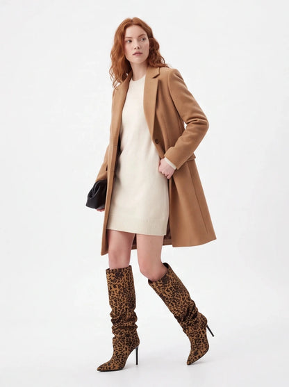 A model wearing a camel mid-length coat over a cream mini dress, paired with leopard-print slouchy knee-high boots and holding a small black clutch, photographed against a plain white studio background.